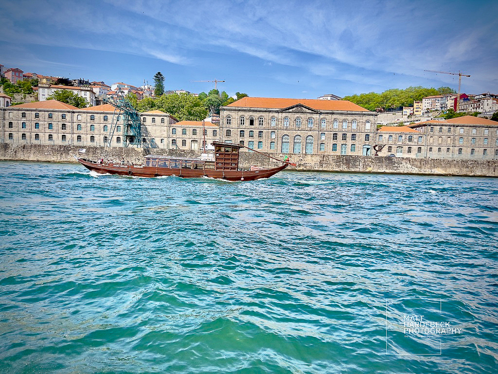Traditional wooden rabelo boat sailing on the Douro River in front of port wine lodges.