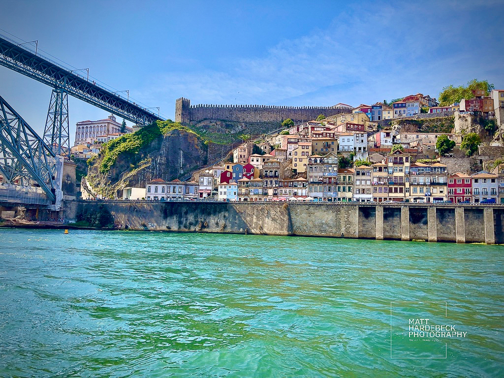 Riverside view of colorful houses in Porto’s Ribeira district beneath the Dom Luís I Bridge, with medieval stone walls above.