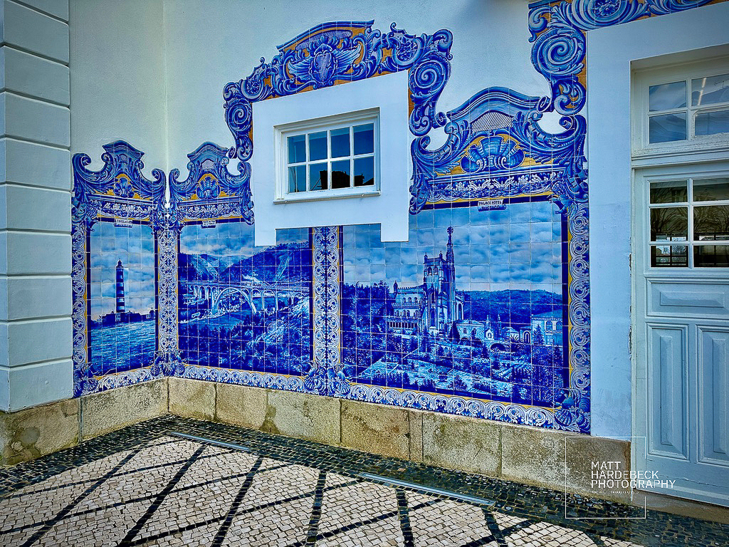 Decorative blue-and-white azulejo tile mural on a white building in Porto, Portugal, depicting local landmarks framed in ornate Baroque-style designs.
