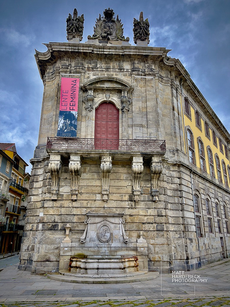 Stone Baroque building with ornate crest, balcony, and a fountain at its base in Porto.