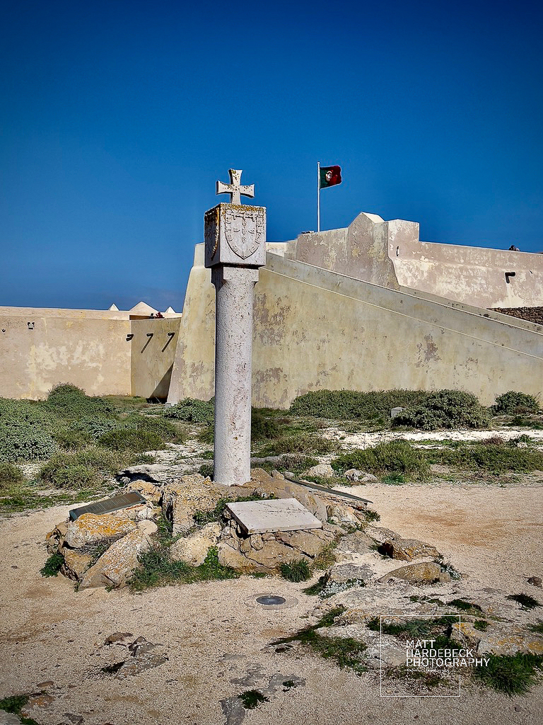 Stone cross monument with Portuguese coat of arms at Fortaleza de Sagres under a clear blue sky.