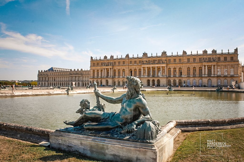 Bronze statue of a reclining river god holding an oar, with Château de Versailles and the pond behind.