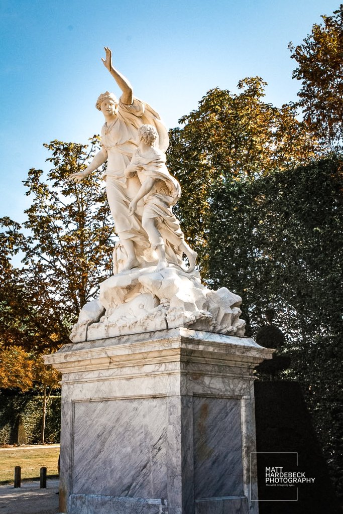 White marble statue of a woman raising her arm while shielding a child, framed by autumn foliage and blue sky.