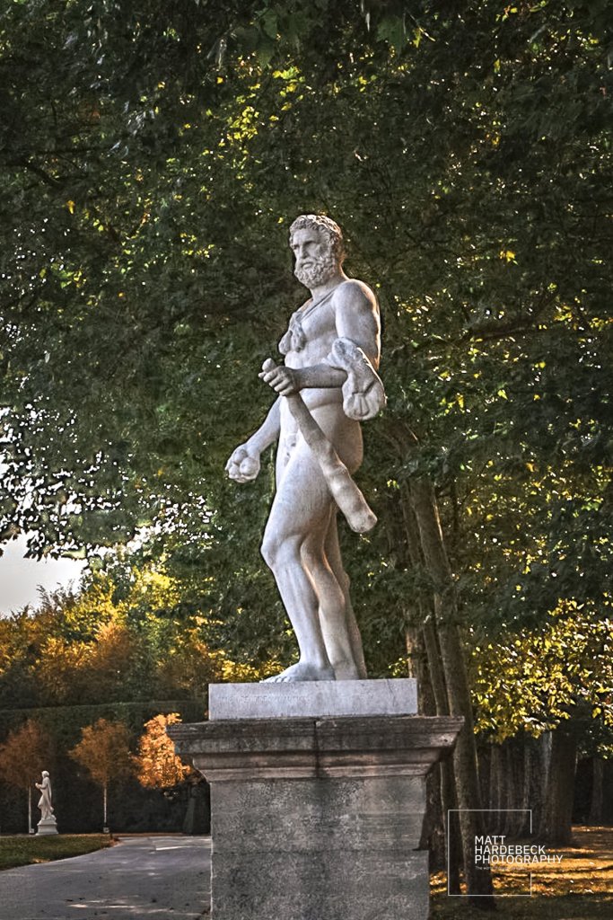 Marble statue of Hercules holding a club and lion skin, partially shaded by tall trees in the gardens of Versailles.