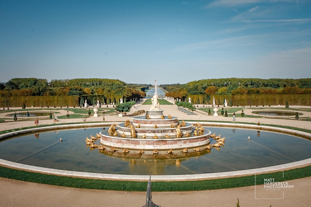 The Latona Fountain at Versailles with golden frogs and marble tiers, framed by the palace gardens and Grand Canal in the distance.