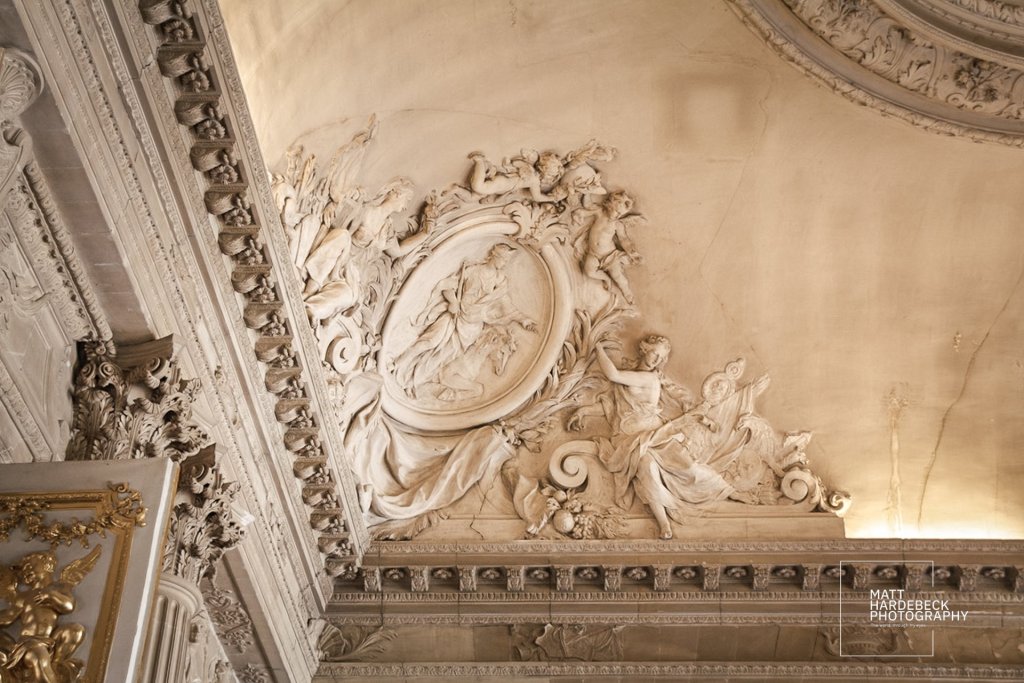 Elaborate plaster relief on the ceiling of a Versailles room, featuring cherubs and classical figures in dynamic poses.