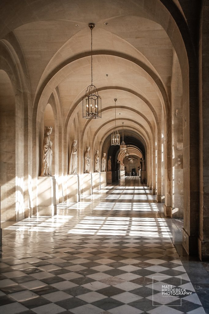 A sunlit arched corridor inside Versailles, lined with statues of queens and a checkered marble floor.