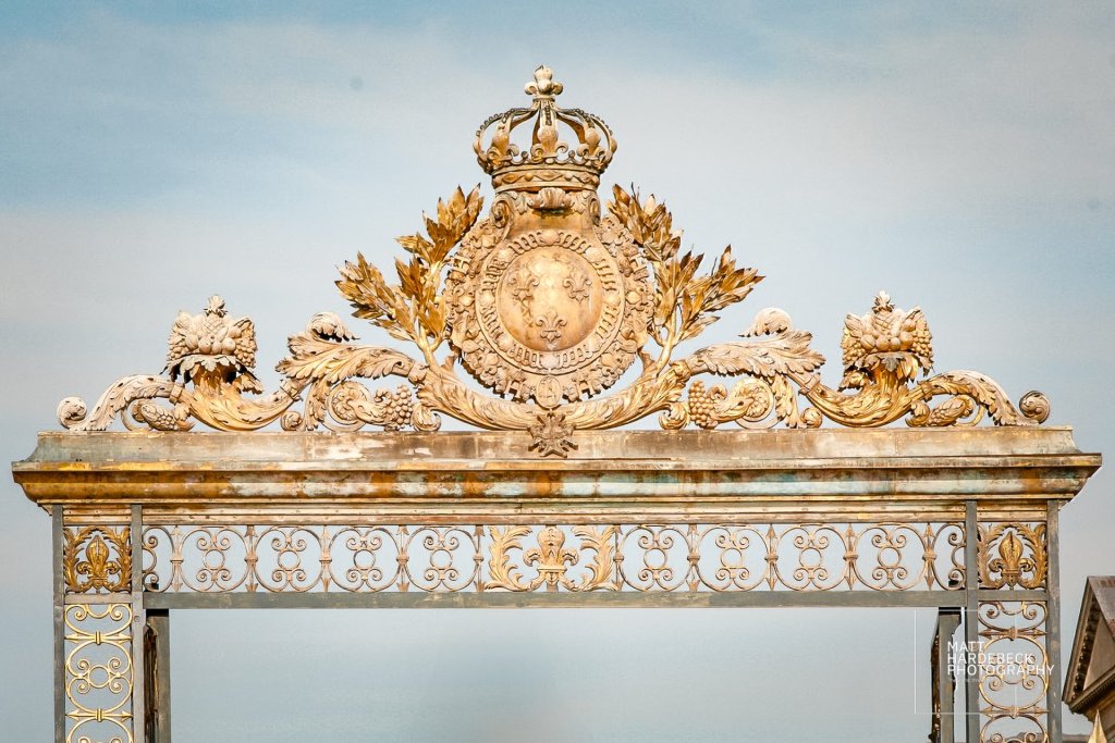 Close-up of the gilded royal crest and fleur-de-lis above the entrance gate to Versailles, with intricate decorative motifs and crown detail.
