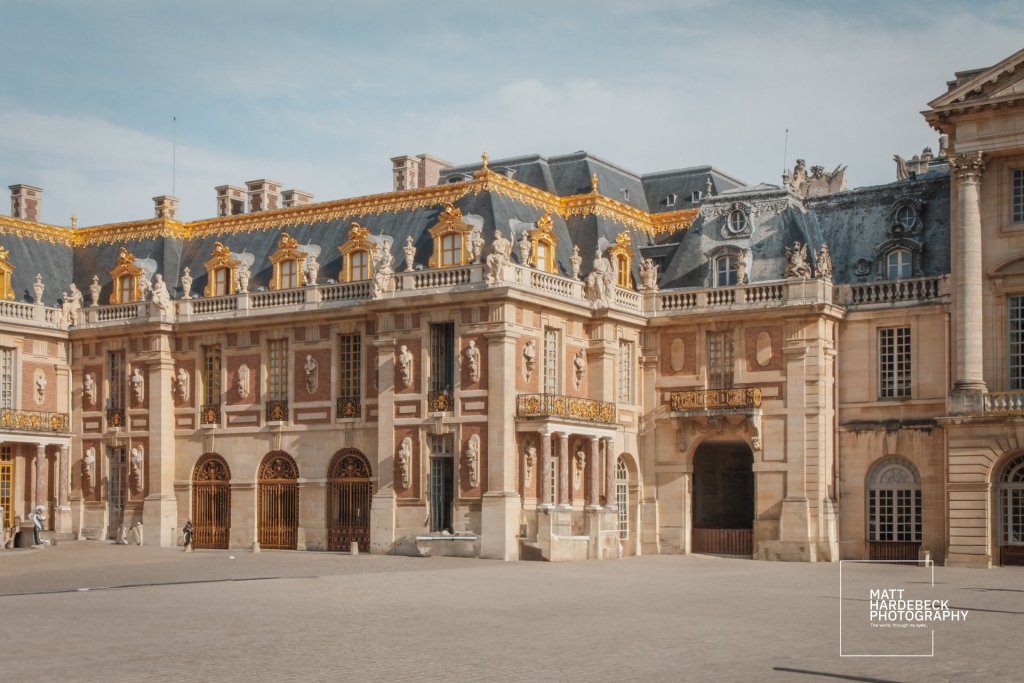 Ornate façade of the central courtyard at the Château de Versailles, featuring gold-trimmed rooflines, arched windows, and classical statues.